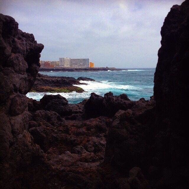 A rocky coastline with waves crashing against the rocks and a building in the background.