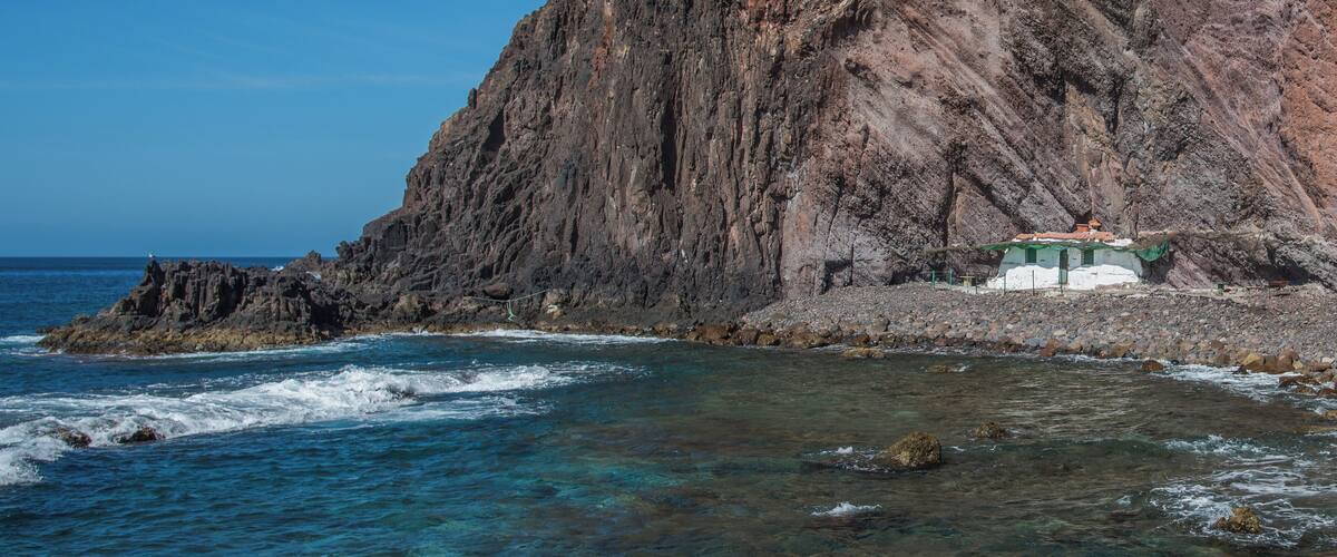 A rocky coastline with clear blue waters, a large cliff, and a small building with a green roof.