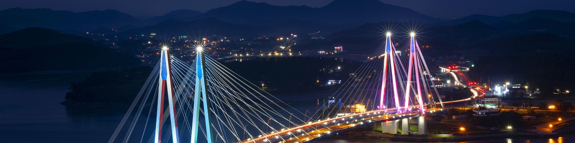 Uma ponte iluminada com vários cabos, um rio e uma paisagem urbana ao fundo.