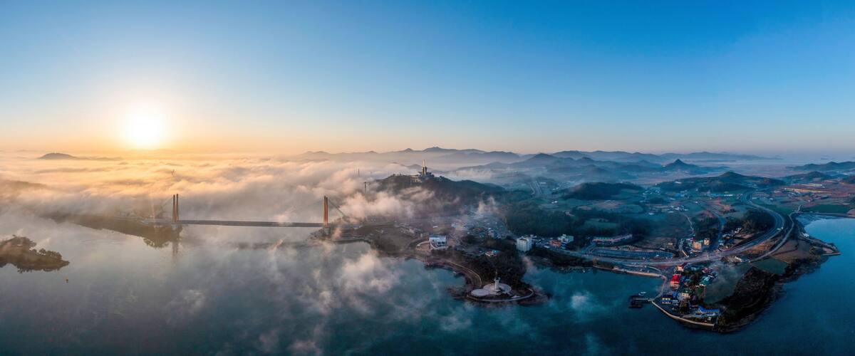 A sunrise over a coastal city with a prominent bridge, surrounded by fog and mountains.