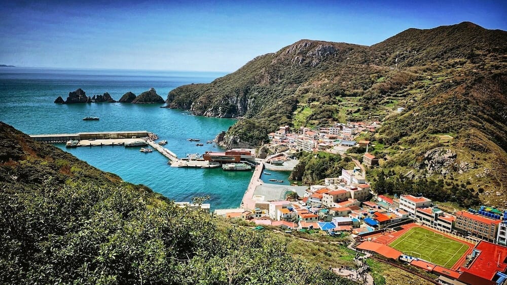 A coastal town with a harbor, rocky cliffs, and a mountainous backdrop.