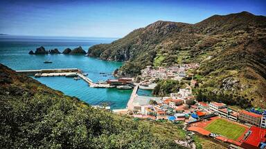 A coastal town with a harbor, rocky cliffs, and a mountainous backdrop.