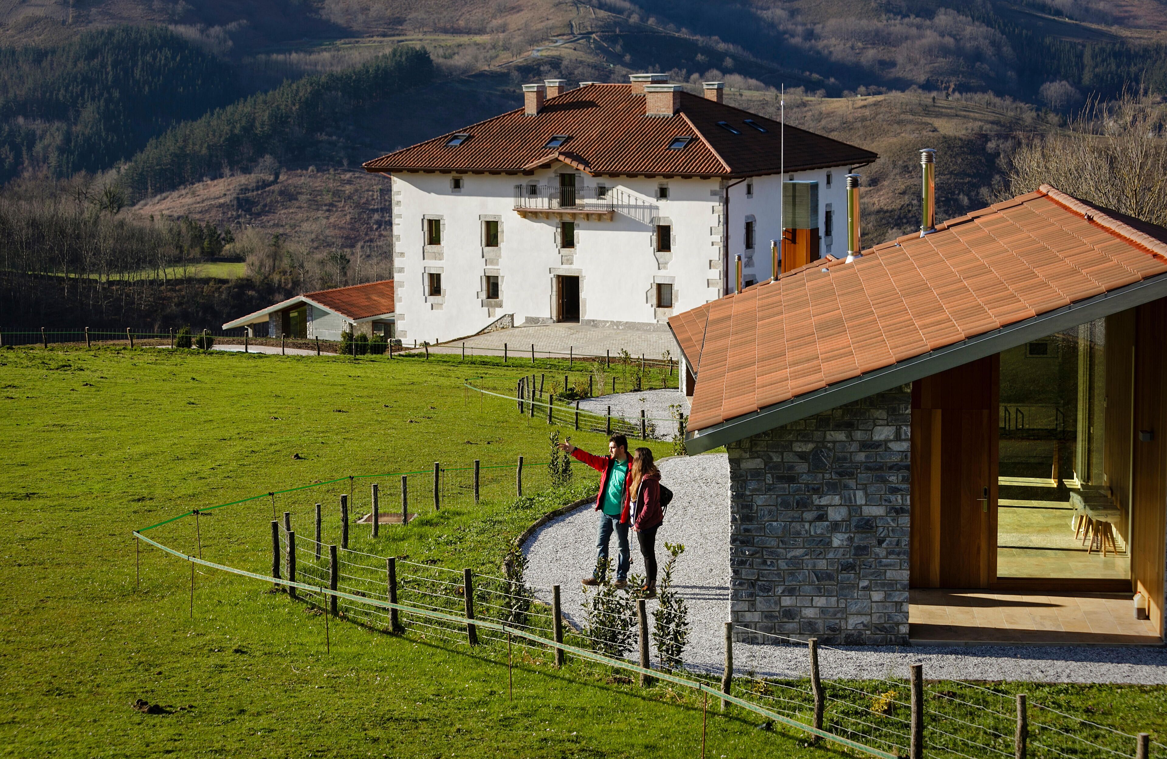 A white house with a red roof, a stone building with a wooden door, and a fenced green field with mountains in the background.