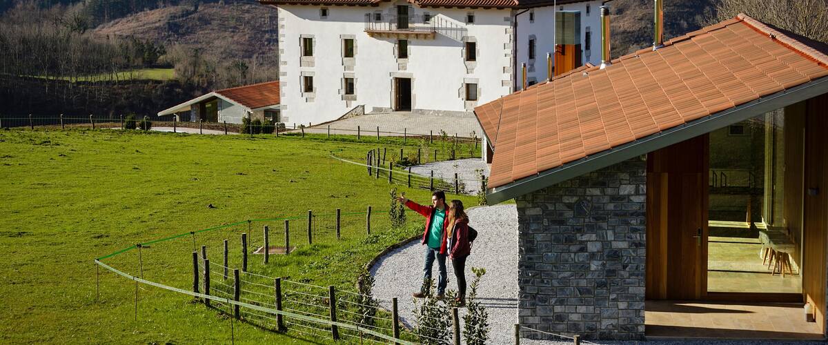 A white house with a red roof, a stone building with a wooden door, and a fenced green field with mountains in the background.