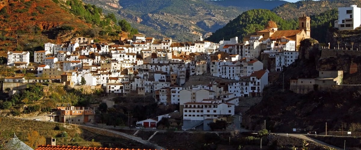 A coastal town with white buildings nestled in a valley, surrounded by hills and mountains.