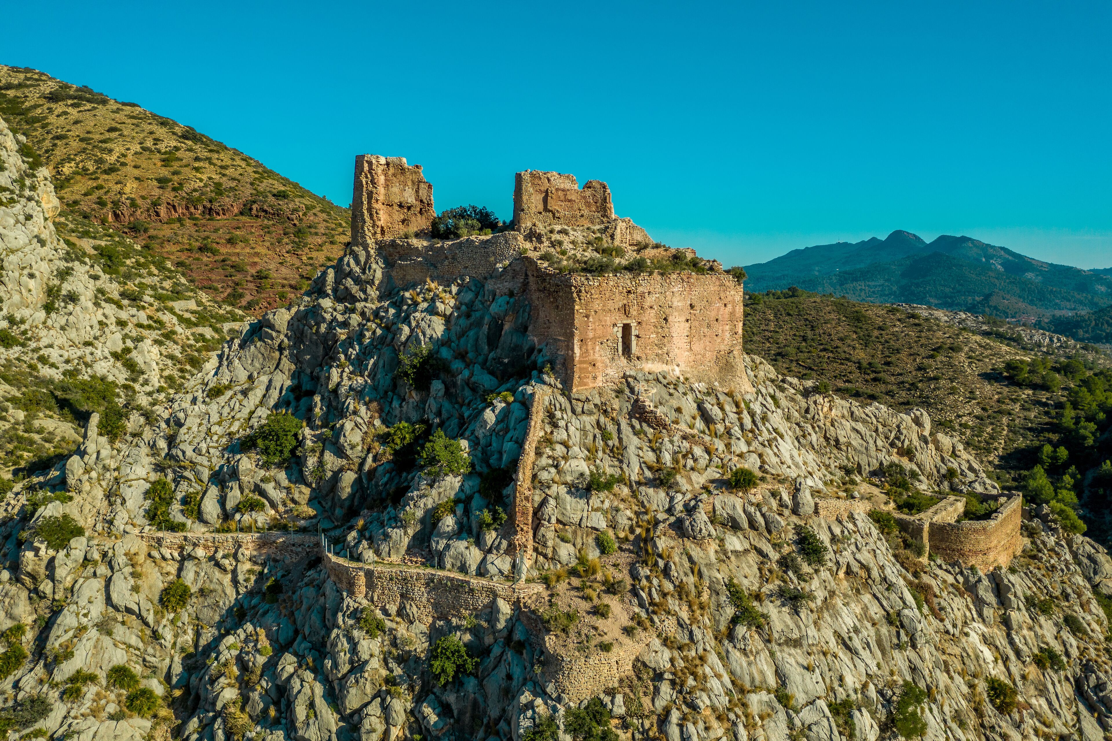 A rocky fortress with ruins on a hill, surrounded by mountains and sparse vegetation.
