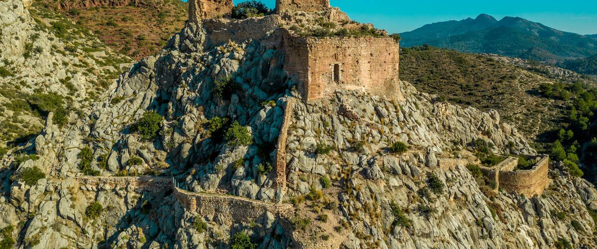 A rocky fortress with ruins on a hill, surrounded by mountains and sparse vegetation.