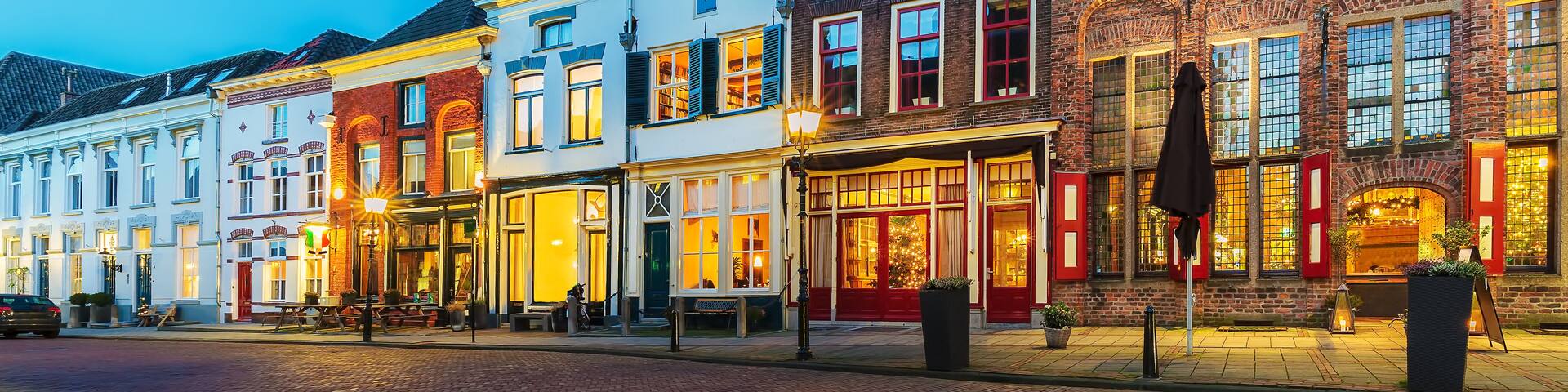 A row of brightly lit historic buildings with distinct architectural styles, a cobblestone street, and a clear evening sky.