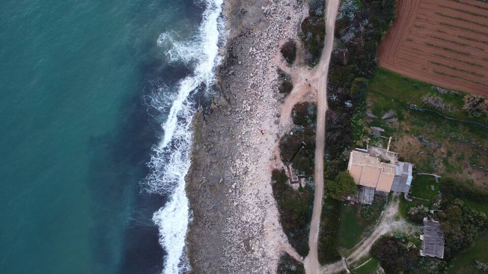 An aerial view of a coastal area with a rocky shoreline, a dirt path, and a few buildings.