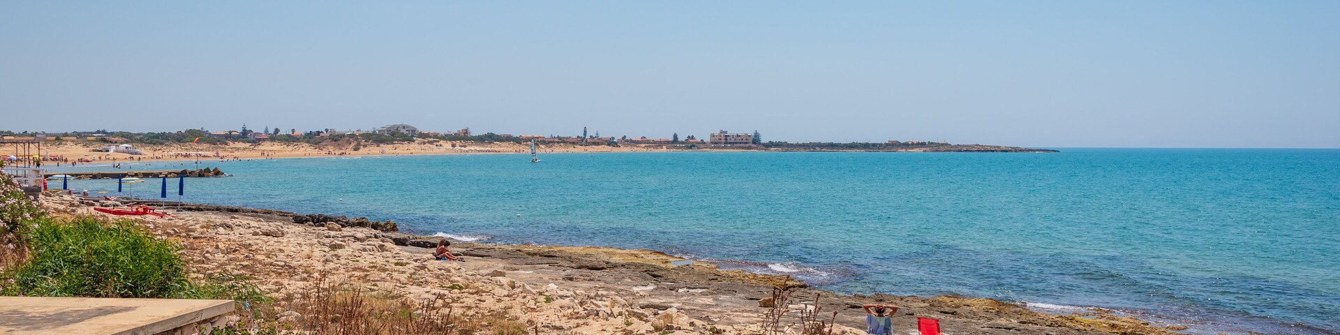 Paisagem costeira com praia rochosa, mar azul cristalino, linha de costa distante com edifícios e um cais que se estende na água.