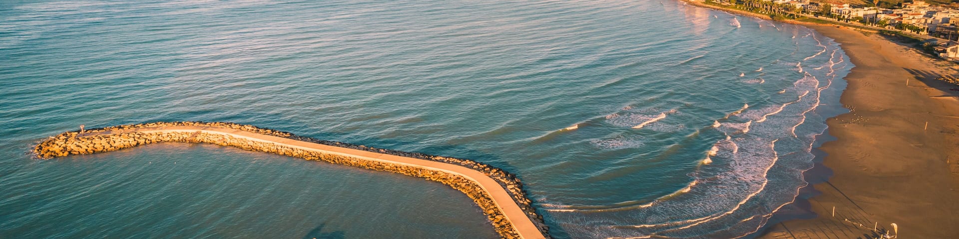 A coastal landscape with a sandy beach, a rocky breakwater, and a distant shoreline with buildings.