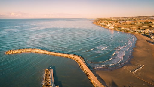 Paisagem costeira com uma praia arenosa, um molhe rochoso e uma linha de costa distante com edifícios.