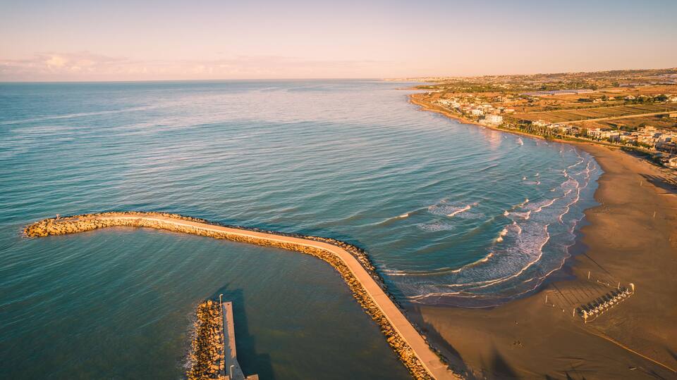A coastal landscape with a sandy beach, a rocky breakwater, and a distant shoreline with buildings.