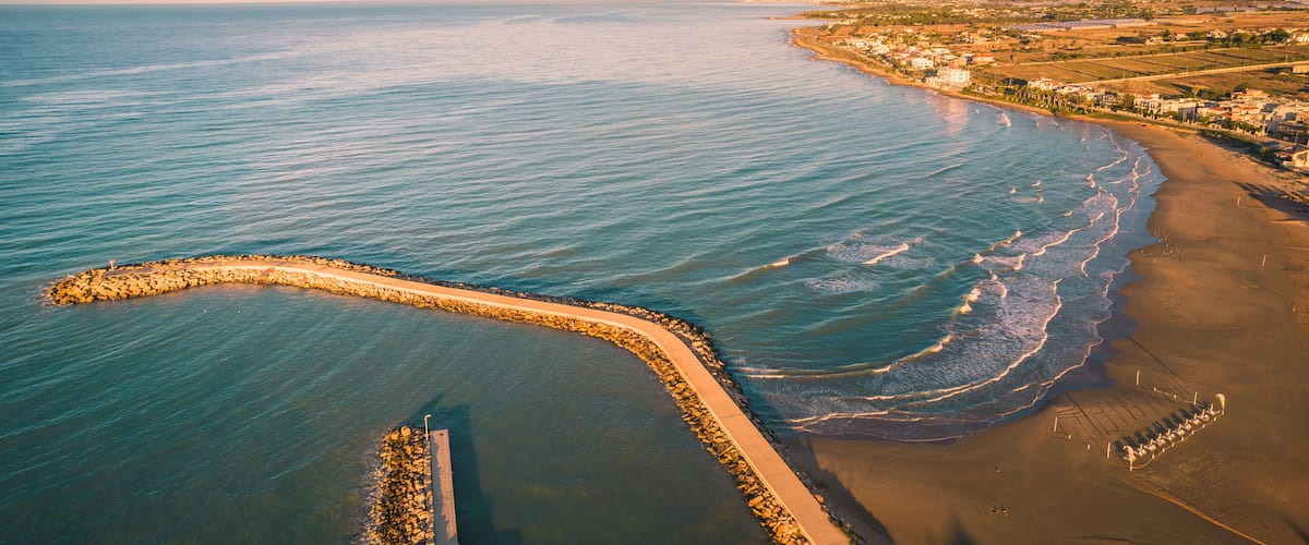 A coastal landscape with a sandy beach, a rocky breakwater, and a distant shoreline with buildings.