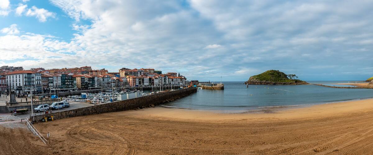 A coastal town with a sandy beach, a rocky island, and a marina with boats.