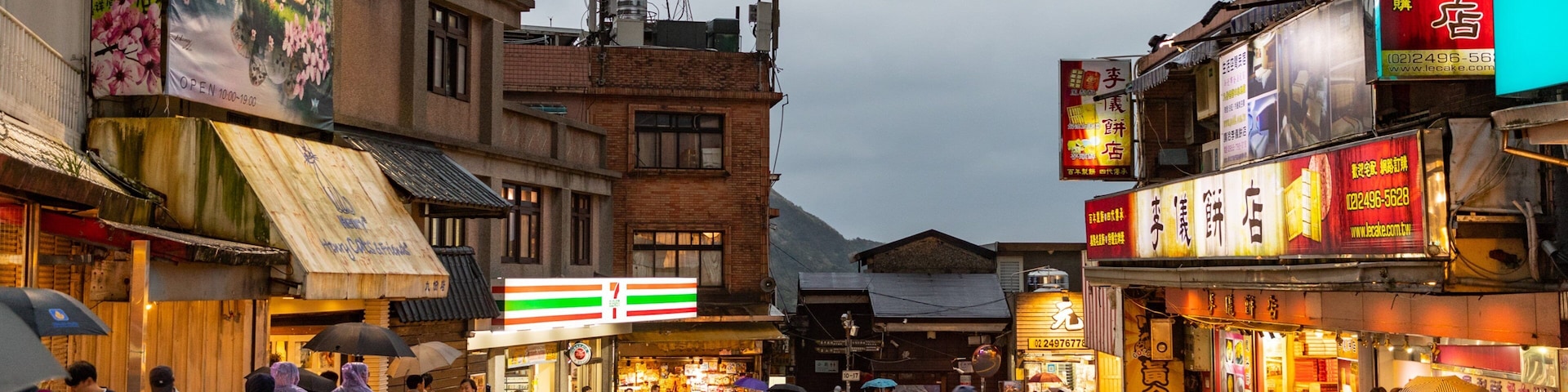 A busy street with shops, a 7-Eleven, and a mountain in the background.