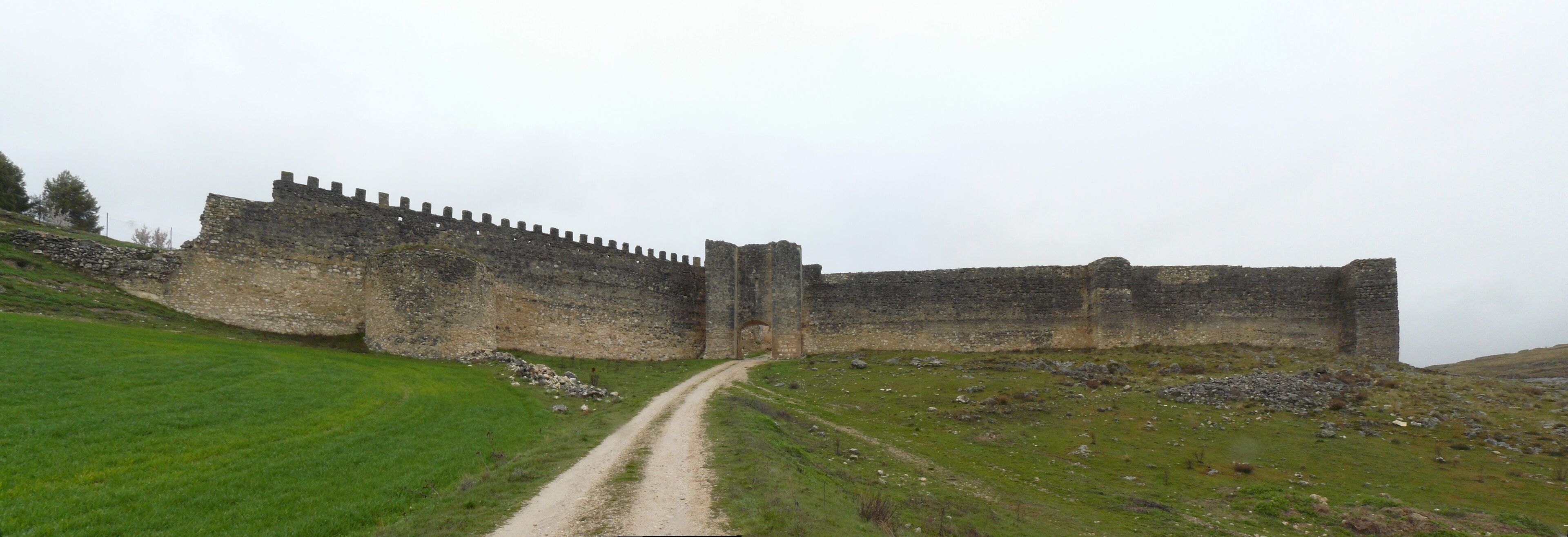 A medieval stone castle with a long, narrow entrance gate, surrounded by grassy hills and a clear sky.