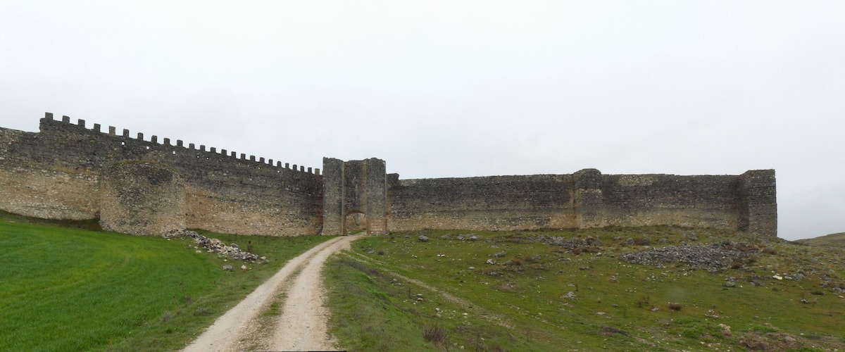 A medieval stone castle with a long, narrow entrance gate, surrounded by grassy hills and a clear sky.