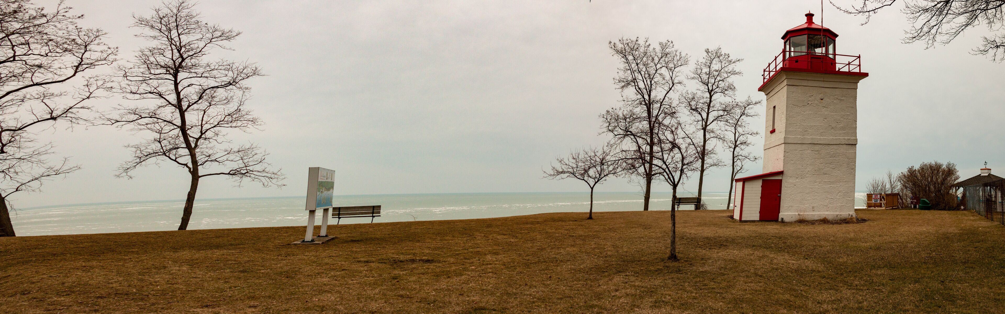 A lighthouse, a bench, and leafless trees by the ocean.