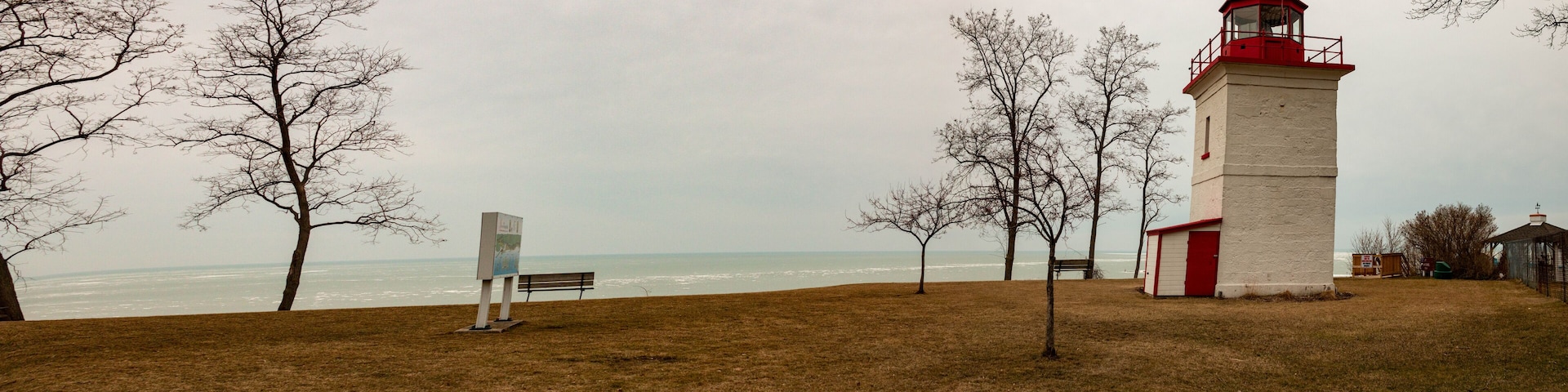 A lighthouse, a bench, and leafless trees by the ocean.