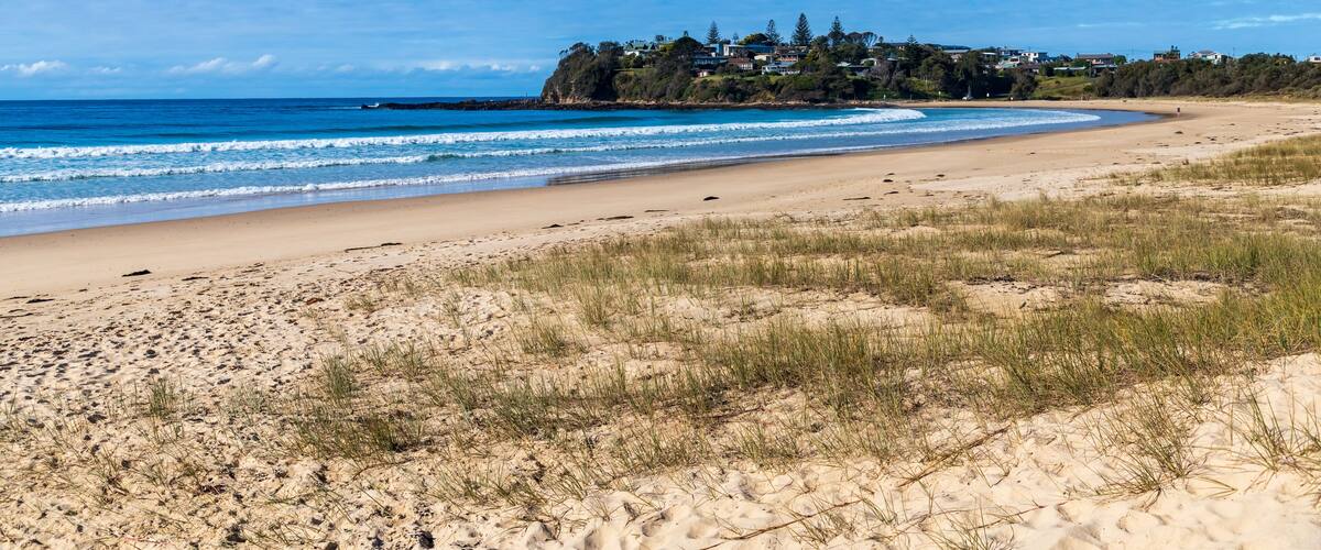 A sandy beach with gentle waves, a clear blue sky, and a coastal town with houses on a cliff.