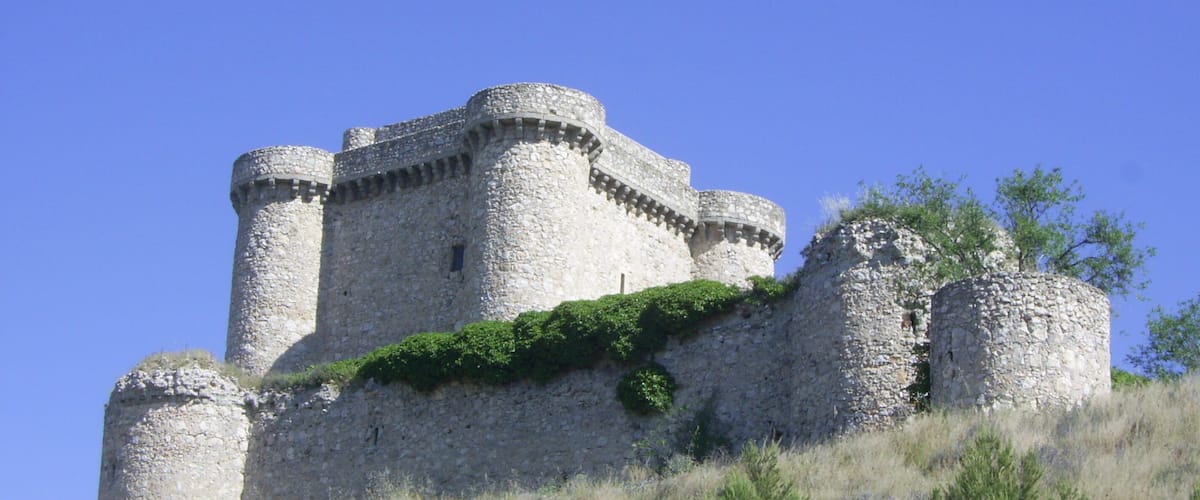 Un castillo medieval con muros y torres de piedra, rodeado de hierba y árboles en una colina.