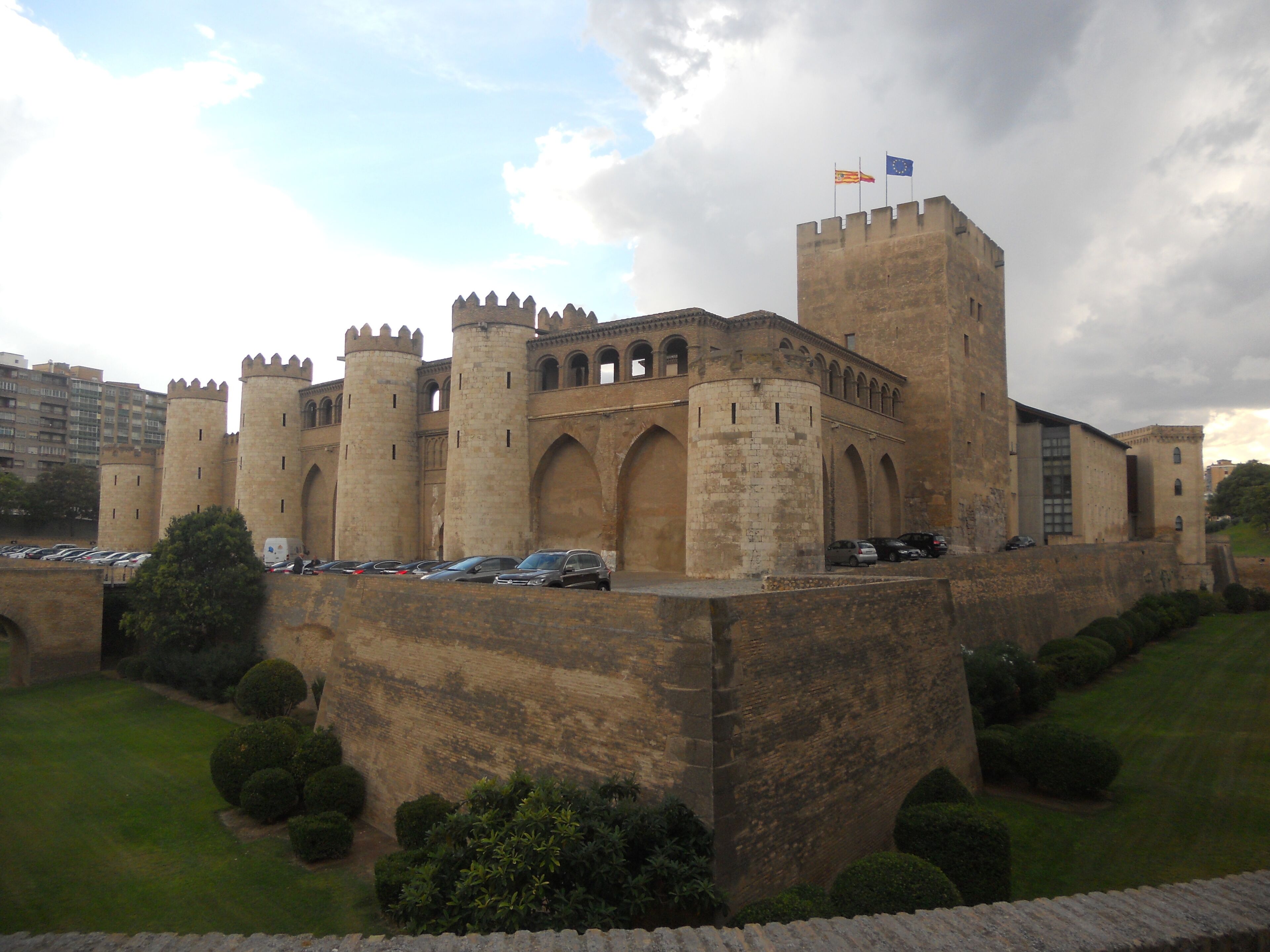 Un château historique doté de nombreuses tours et de fenêtres en arc de cercle, entouré d’une pelouse bien entretenue et d’un espace de stationnement.