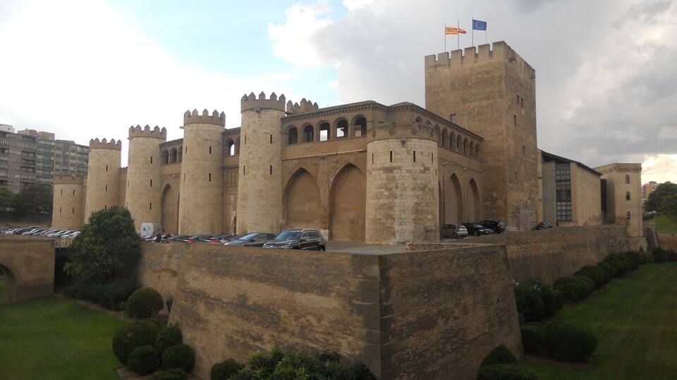 Un château historique doté de nombreuses tours et de fenêtres en arc de cercle, entouré d’une pelouse bien entretenue et d’un espace de stationnement.