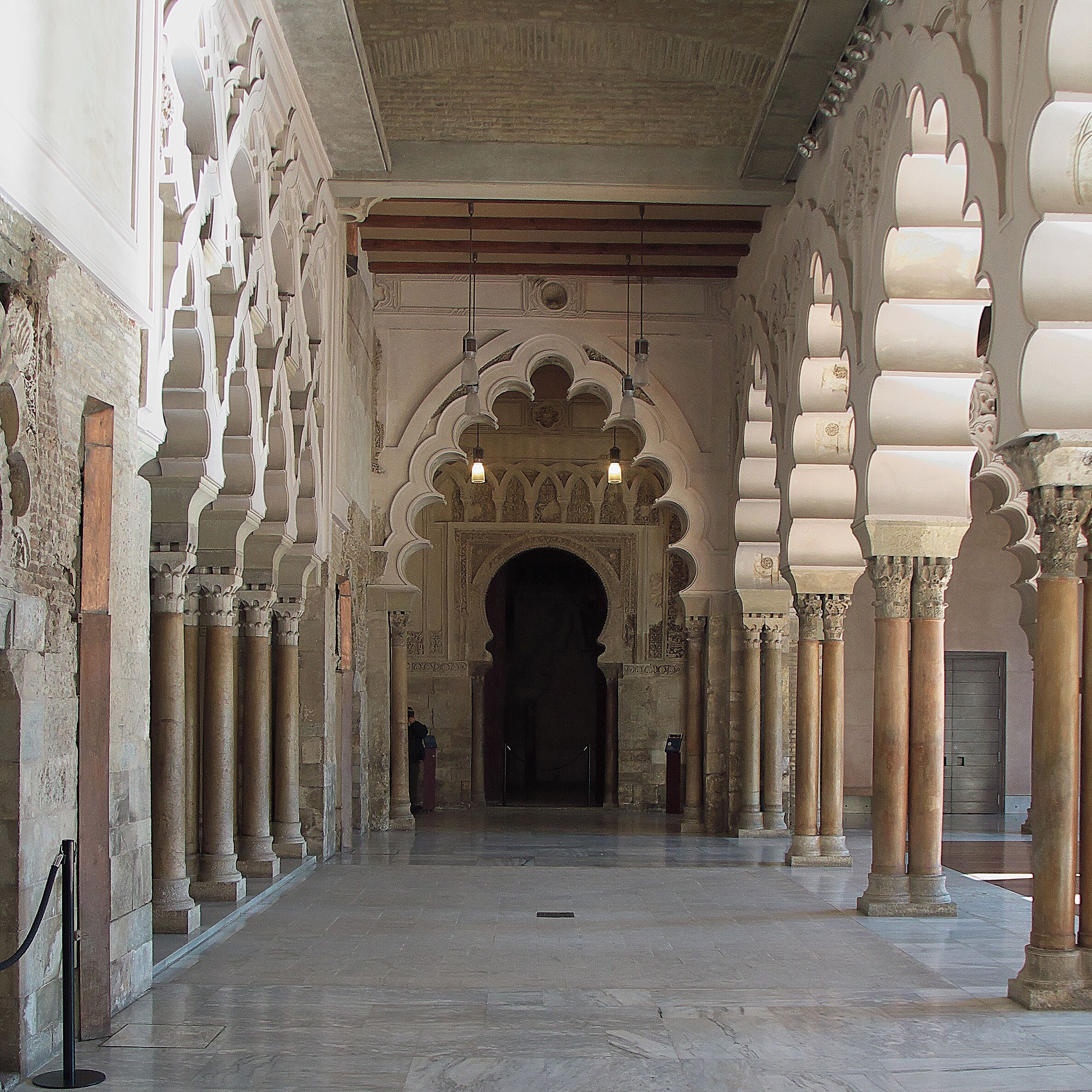 Un couloir intérieur, orné d’arches et de colonnes, qui mène à une porte sombre.