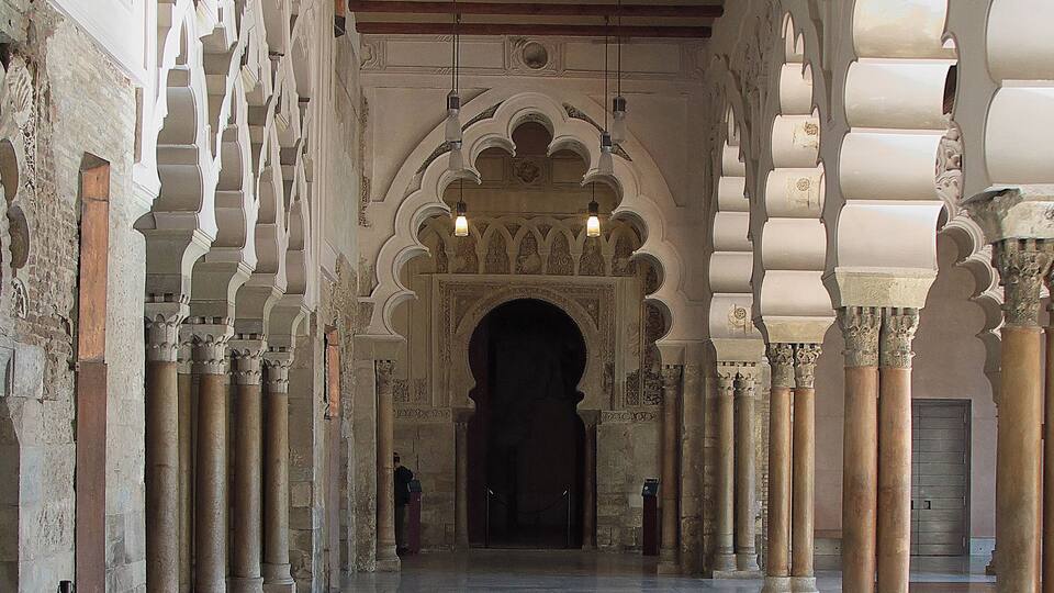 Un couloir intérieur, orné d’arches et de colonnes, qui mène à une porte sombre.