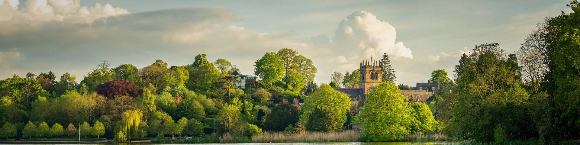 A calm lake reflecting trees and a church with a tall spire.