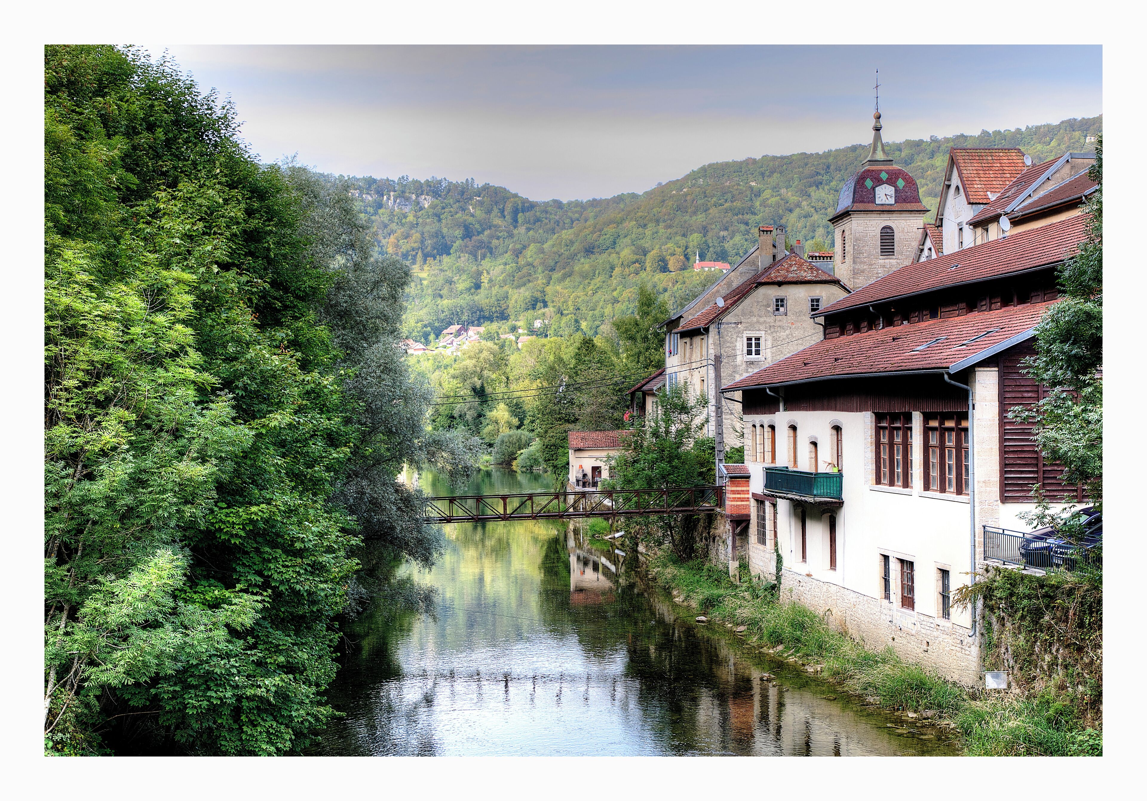 A river flowing through a town with buildings on the right and a forested hillside in the background.