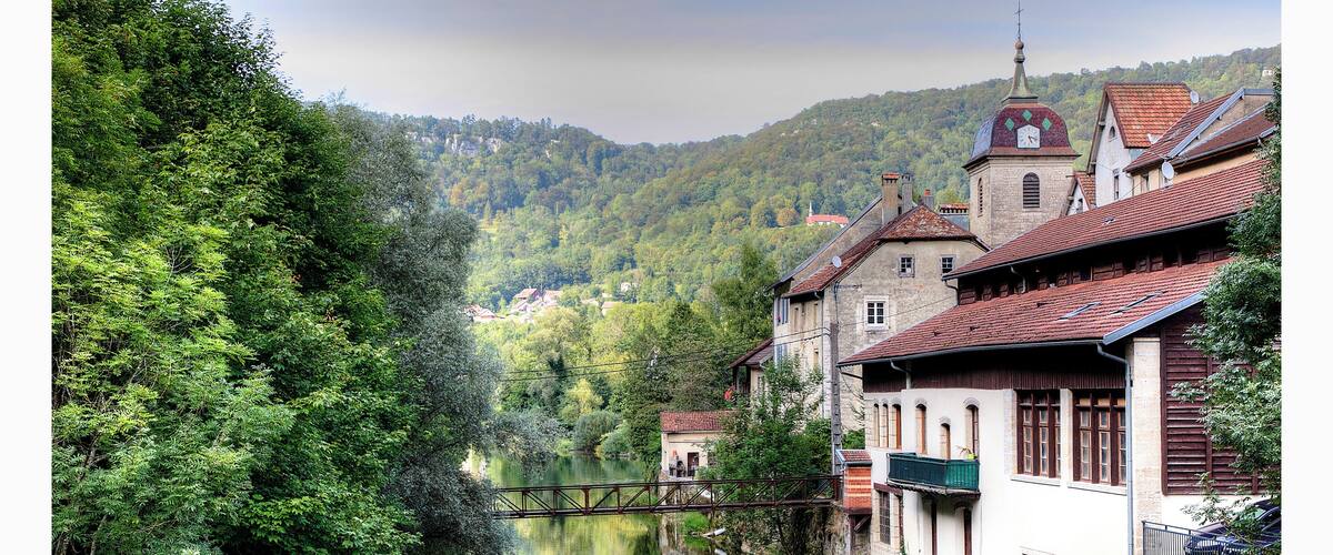 A river flowing through a town with buildings on the right and a forested hillside in the background.