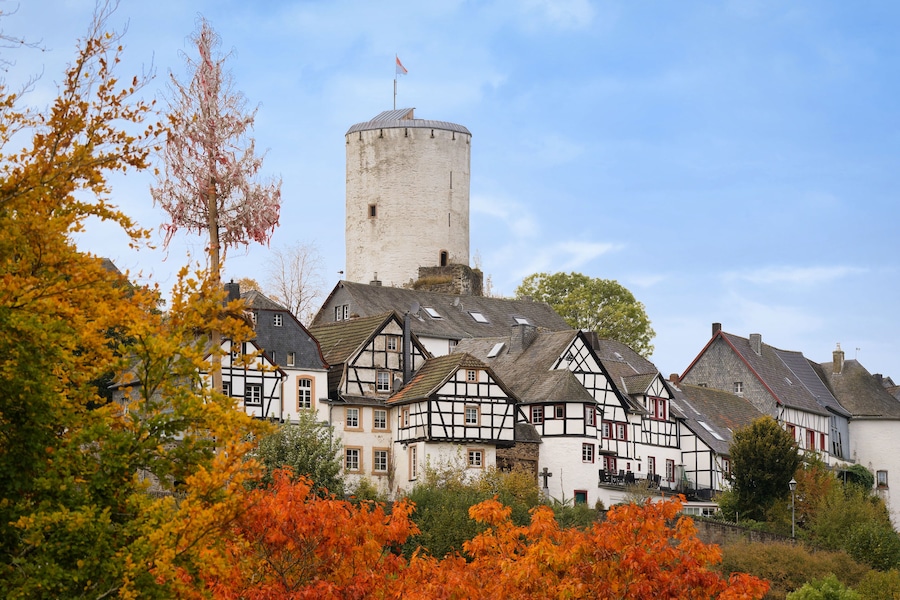 Historic view of Reifferscheid with white castle tower and traditional half-timbered houses. Autumn foliage and clear sky, Eifel region, Germany