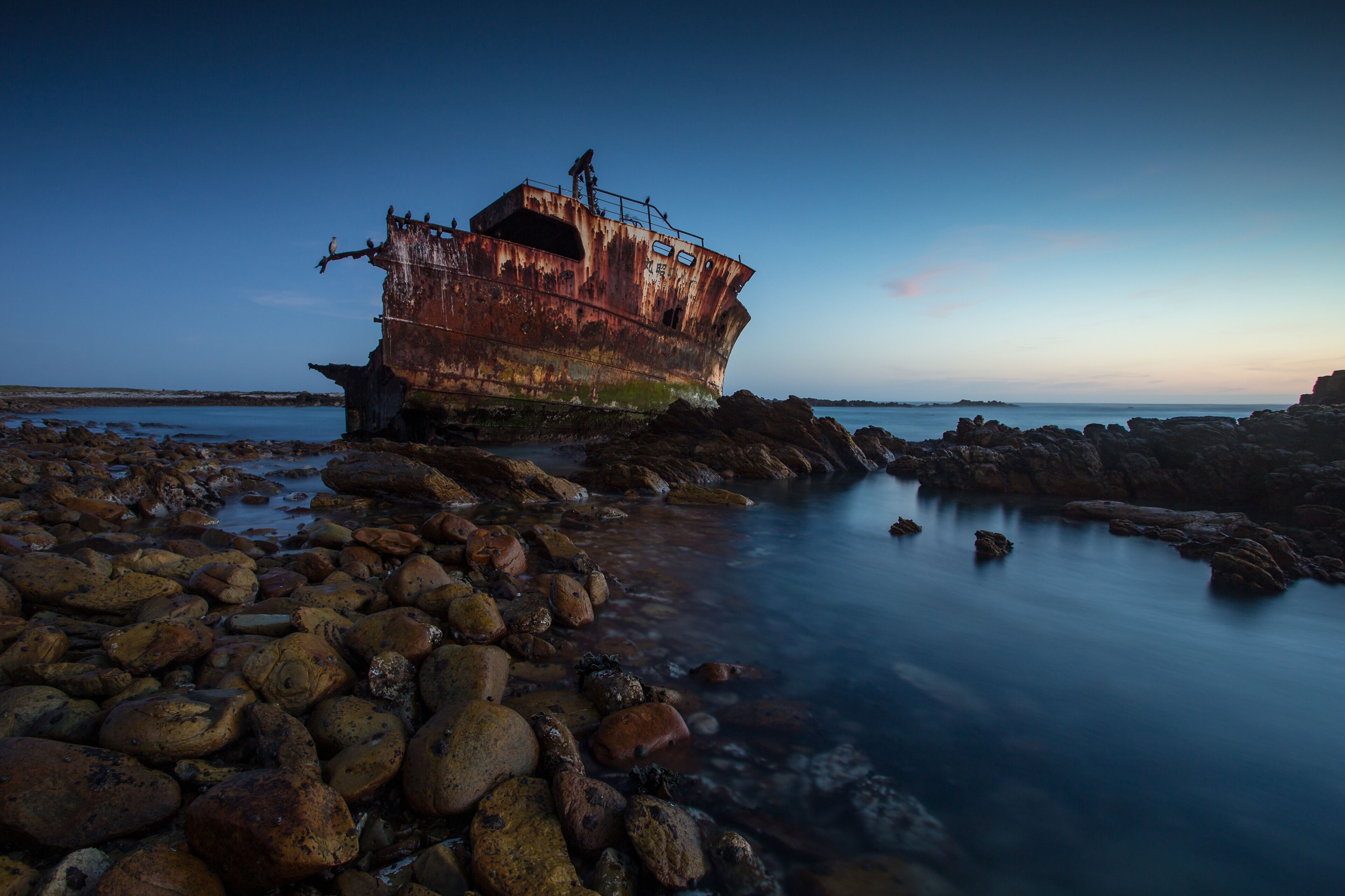 A rusted shipwreck on a rocky shore with calm water.