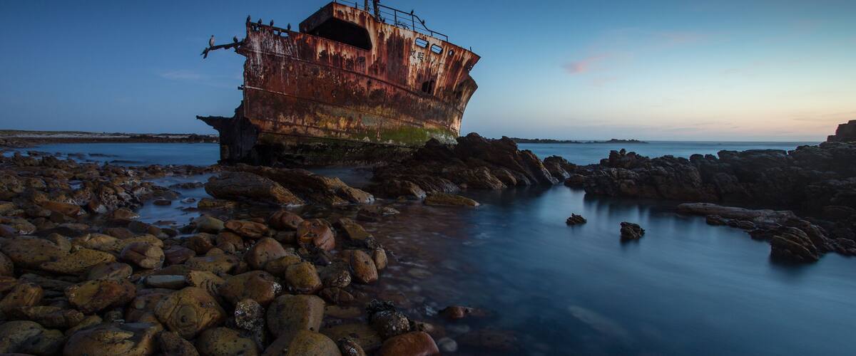 A rusted shipwreck on a rocky shore with calm water.