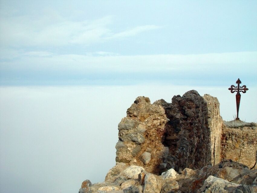 A rocky ruin with a cross on top, overlooking a calm body of water.