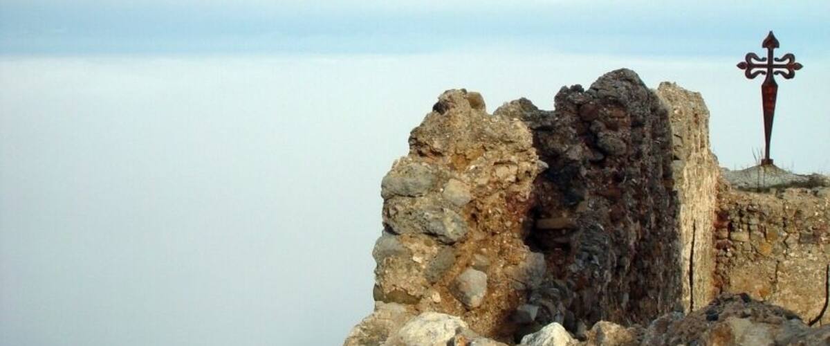 A rocky ruin with a cross on top, overlooking a calm body of water.