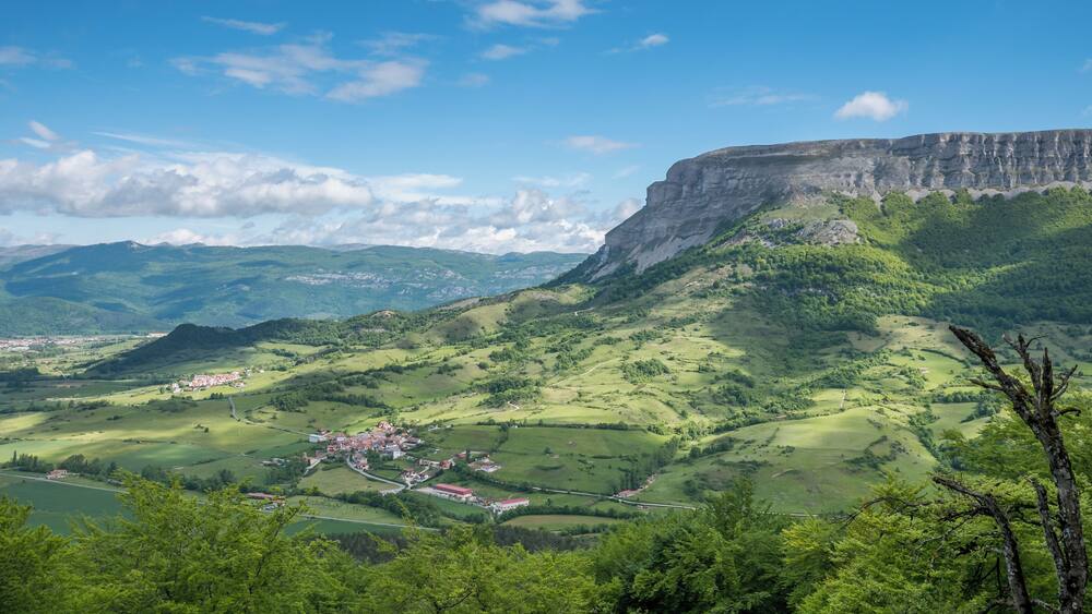 Eine Berglandschaft mit einem Dorf, eingebettet in ein Tal, umgeben von üppiger Vegetation und einer markanten Klippe.