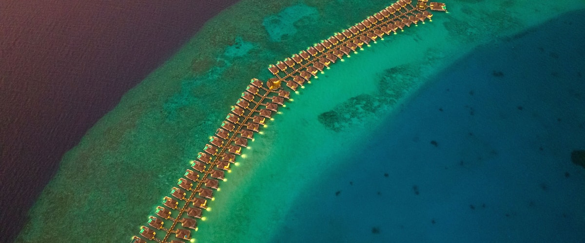 An aerial view of a long, illuminated resort on a crescent-shaped island surrounded by clear turquoise waters.