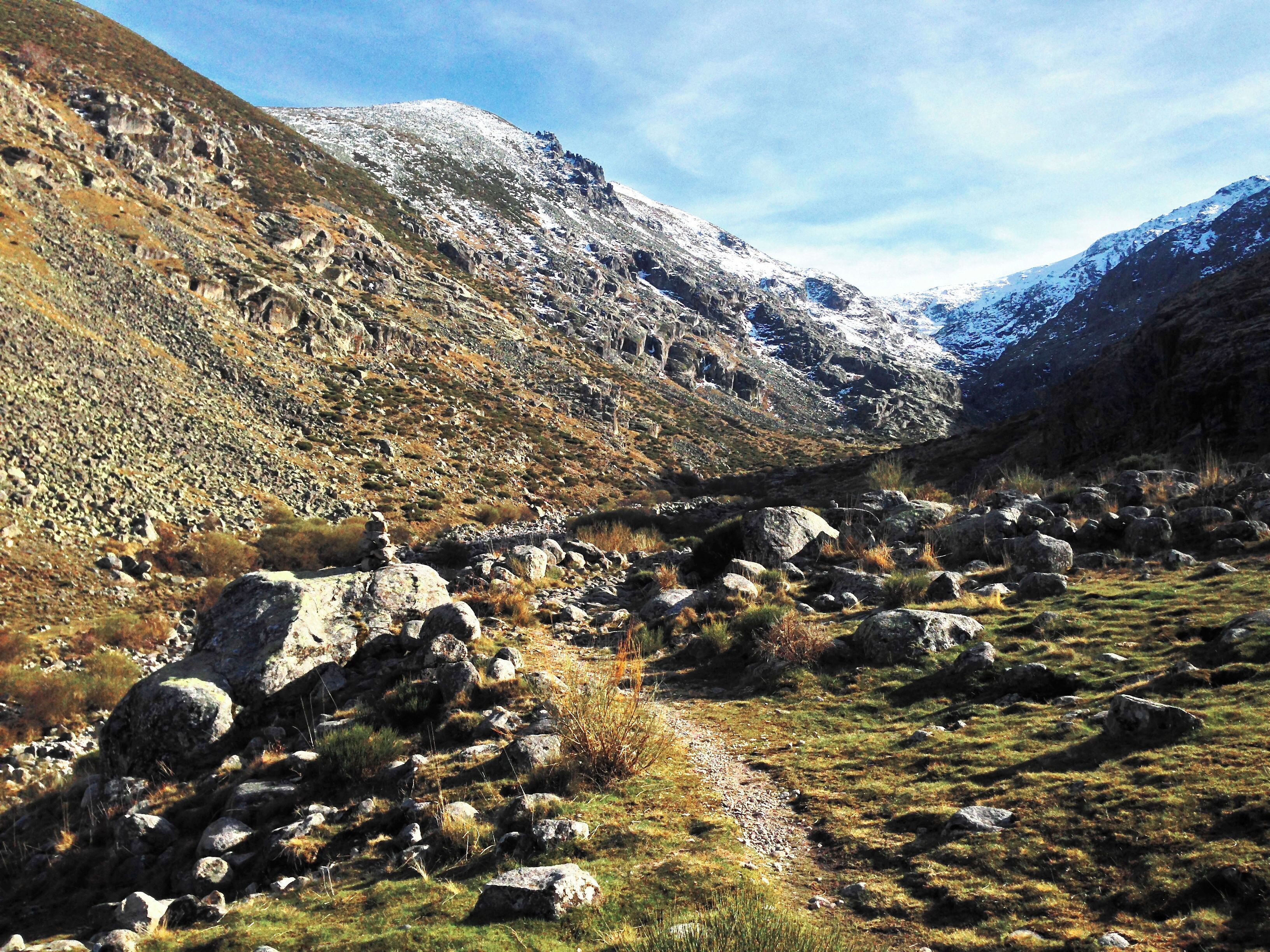 A mountainous landscape with a rocky path, sparse vegetation, and snow-capped peaks under a clear sky.