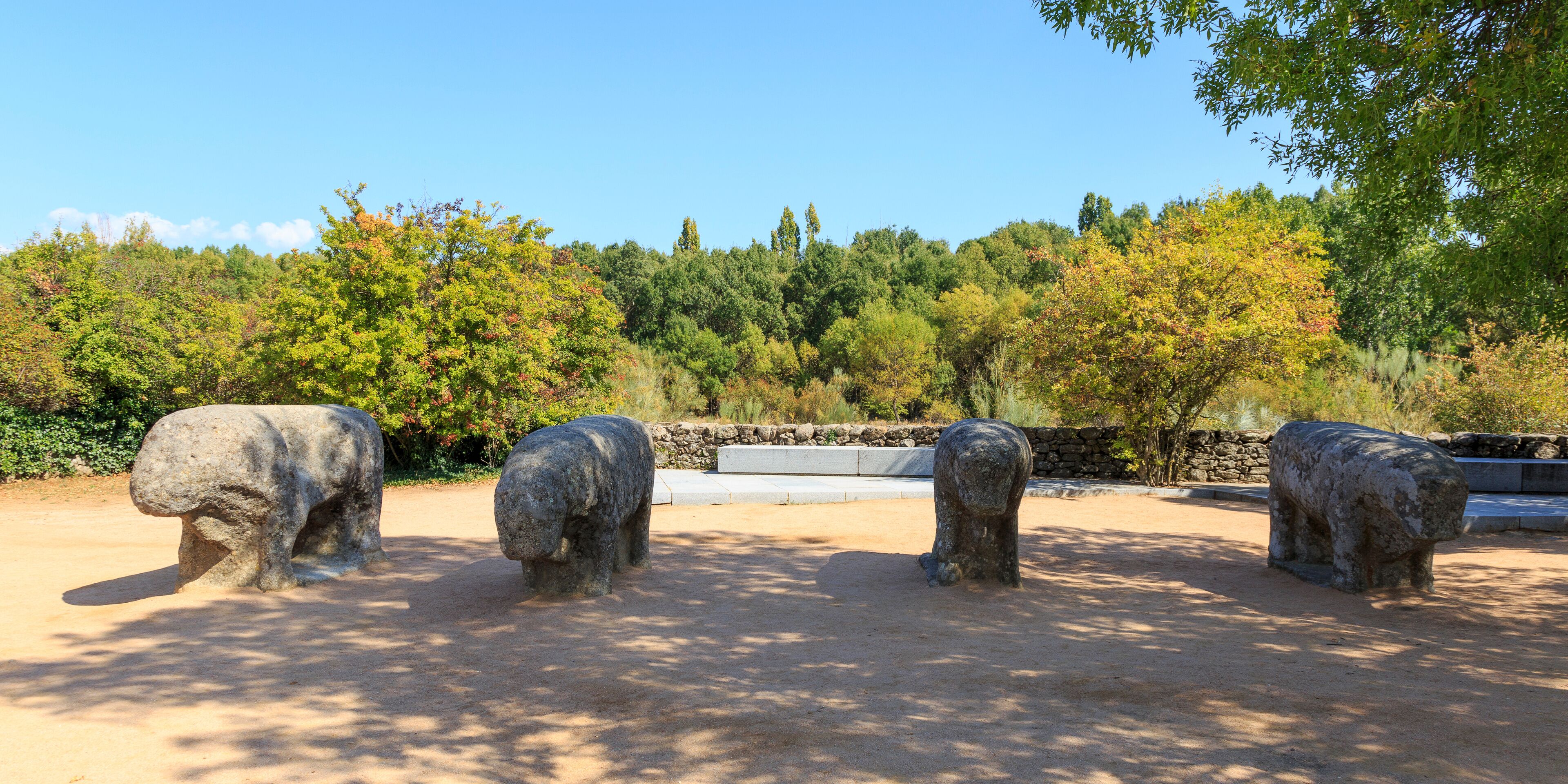 Three stone elephant sculptures in a park with trees and a stone wall in the background.