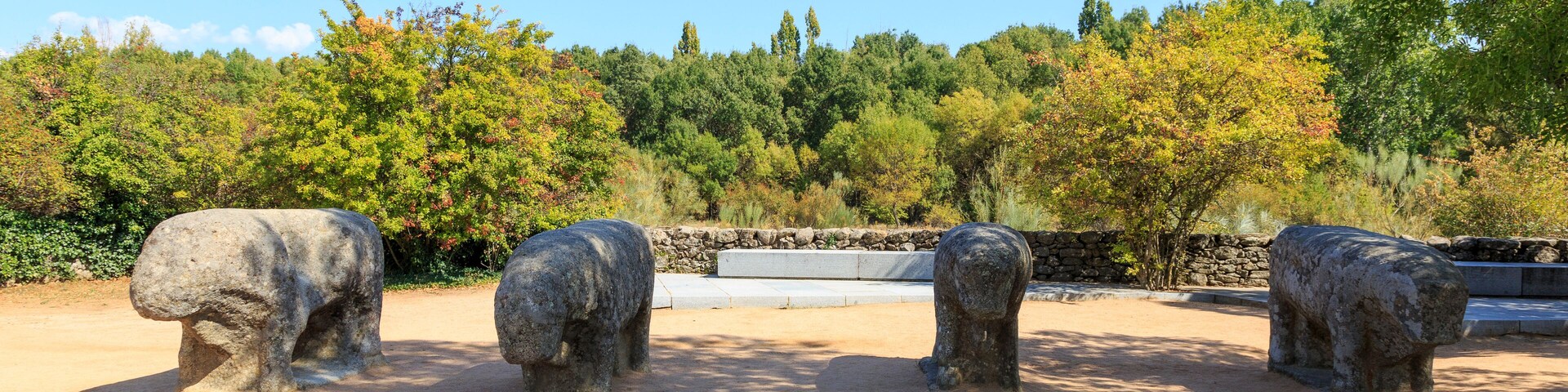 Three stone elephant sculptures in a park with trees and a stone wall in the background.