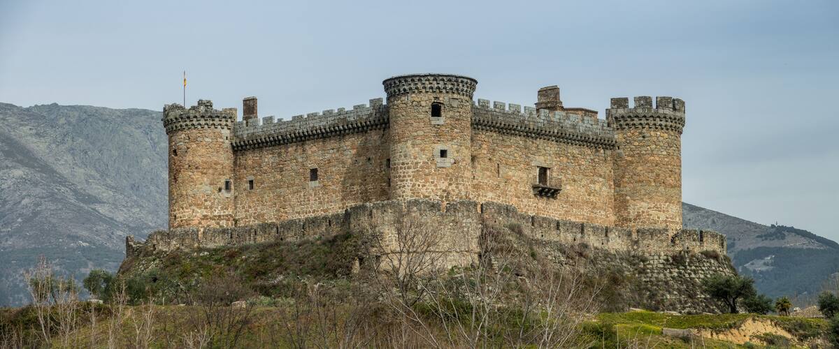 A medieval castle with multiple towers and a stone wall, surrounded by a mountainous landscape.