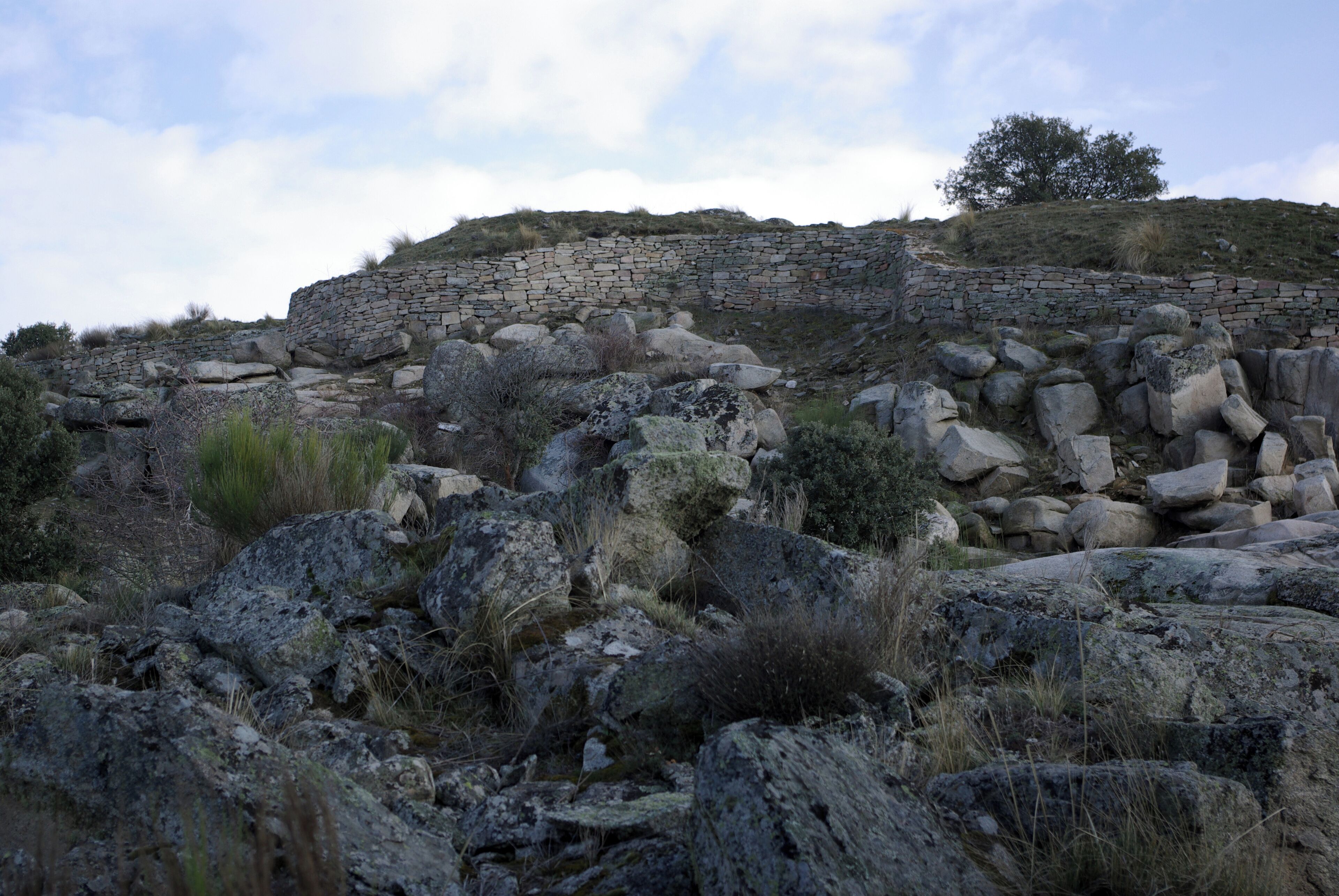 A stone wall on a hillside with scattered rocks and sparse vegetation.