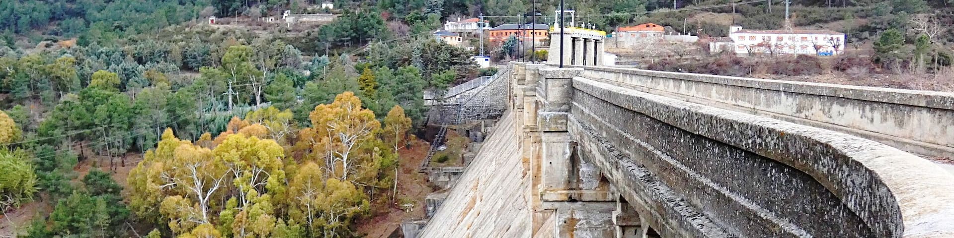 A large dam with a concrete structure, surrounded by a forested landscape and hills in the background.