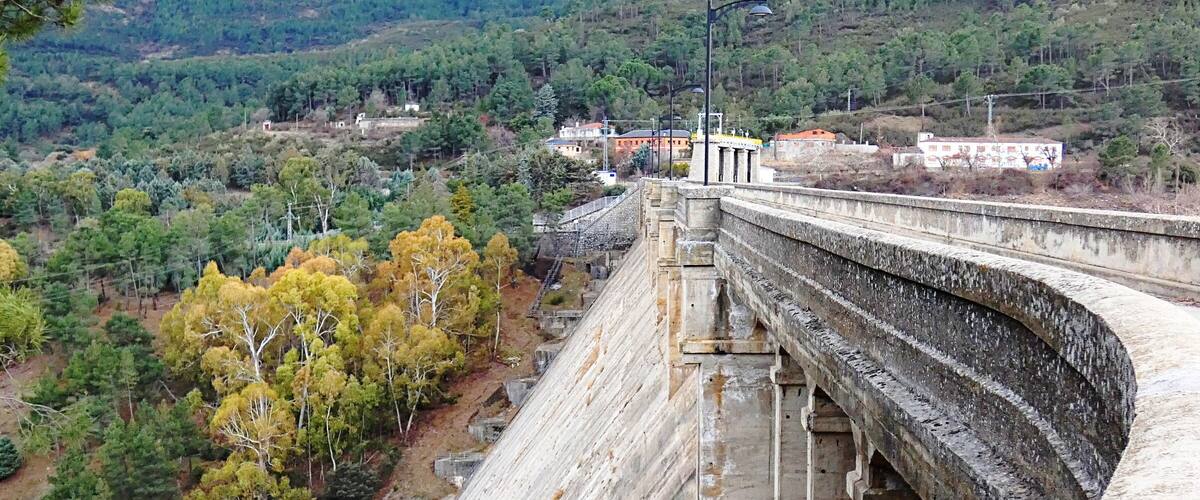 A large dam with a concrete structure, surrounded by a forested landscape and hills in the background.