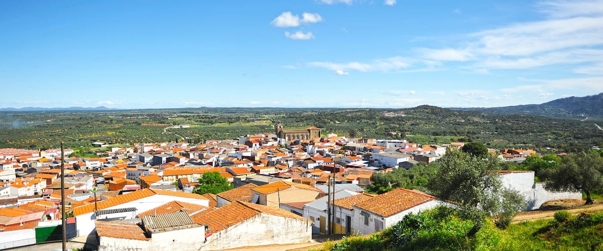 A town with red-tiled roofs, surrounded by green hills and a clear blue sky.