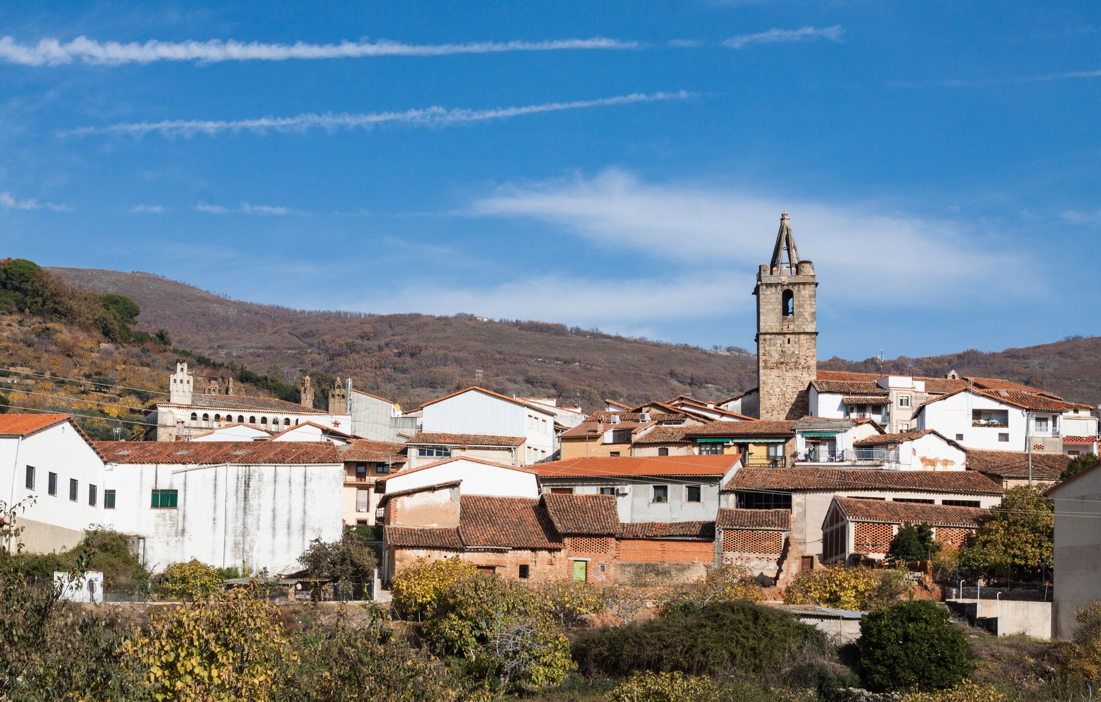 A town with a prominent church tower, white buildings with red-tiled roofs, and a hilly backdrop.