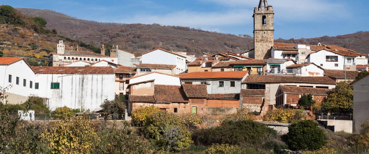 A town with a prominent church tower, white buildings with red-tiled roofs, and a hilly backdrop.